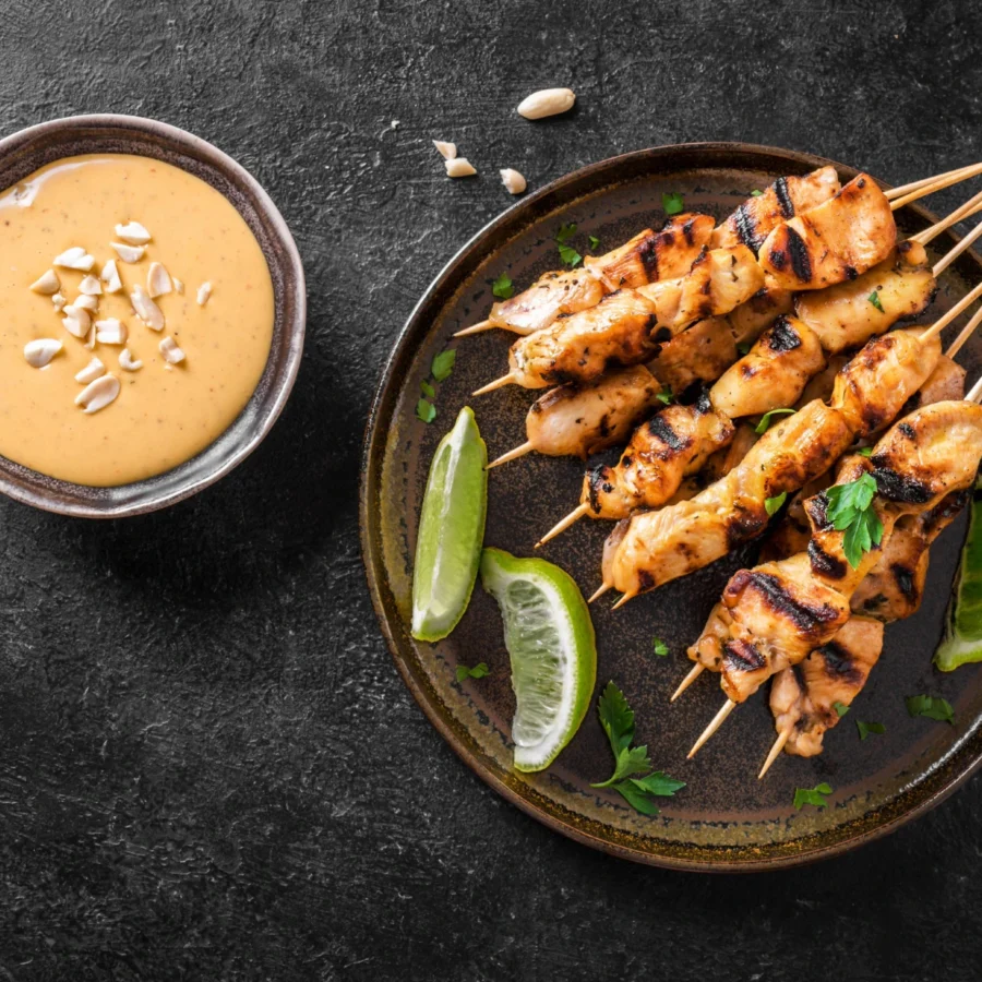 A black plate with chicken satay skewers and dipping sauce.